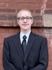Portrait of Elijah Erickson wearing a dark suit and tie in front of a brick wall.